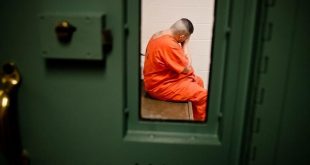 A male prisoner in an orange jump suit is pictured through a green prison door as he sits alone in a cell.