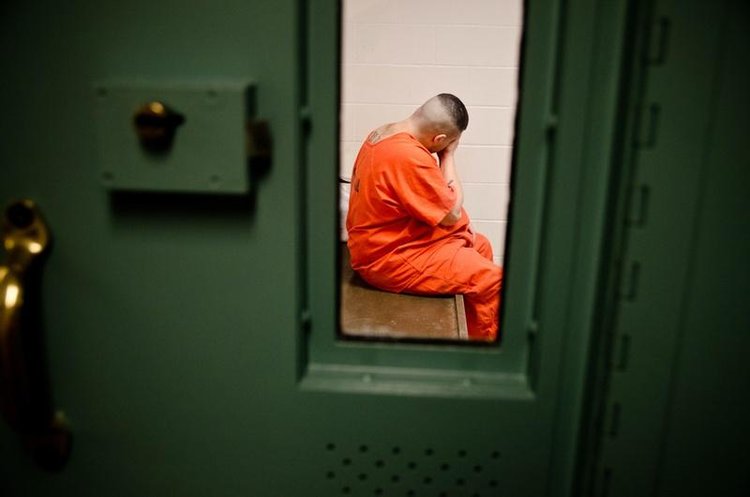 A male prisoner in an orange jump suit is pictured through a green prison door as he sits alone in a cell.