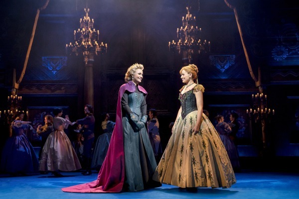 Photo of Samantha Barks as Elsa and Stephanie McKeon as Anna standing and smiling at each other with the Ensemble behind them