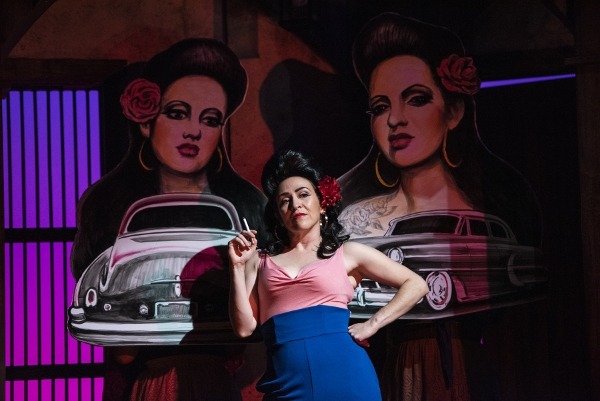 Photo of Sarita Ocón as Dulcinea, standing near a wall on stage and smoking a cigarette