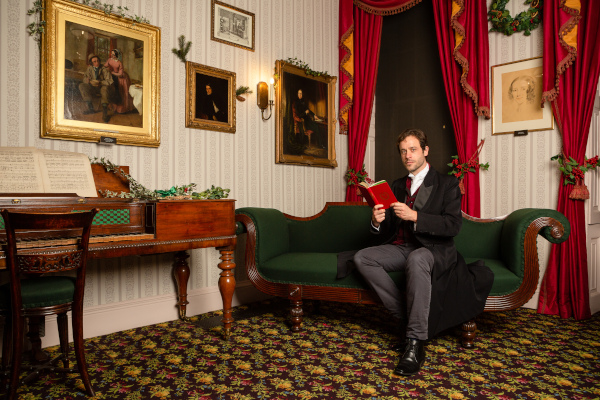 Photo of Dominic Gerrard dressed in a suit and sitting in the Charles Dickens Museum with a book in his hands