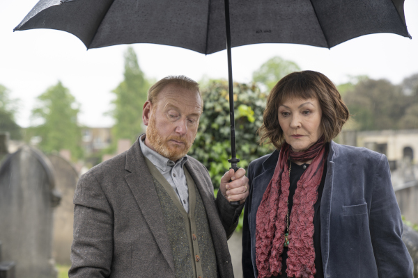 A man and woman standing in a graveyard and sharing an umbrella