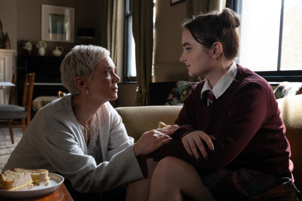 a woman in a white sweater kneeling by a couch and comforting her teenage daughter who is in a maroon school uniform